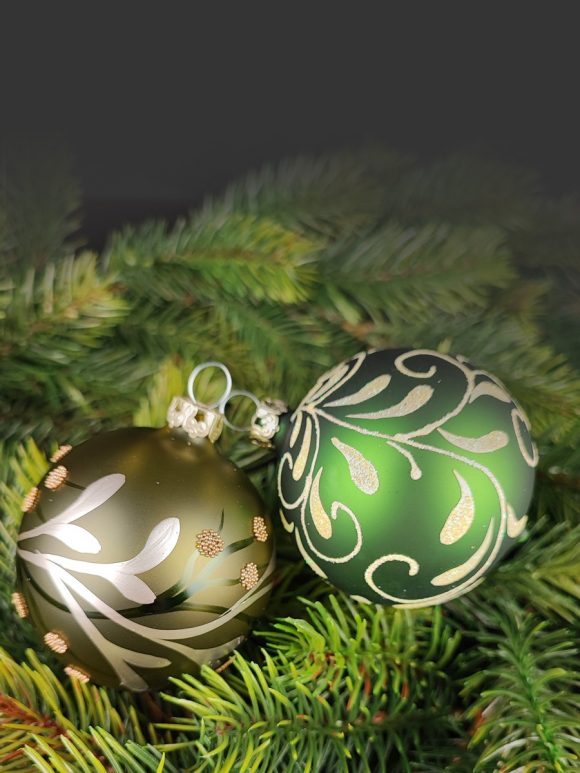 2 Christbaumkugeln aus Glas liegen dekorativ auf grünen Zweigen in einem matten Grün und floralem Design sowie in einem transparentem Khaki-Grün und einer Ranke