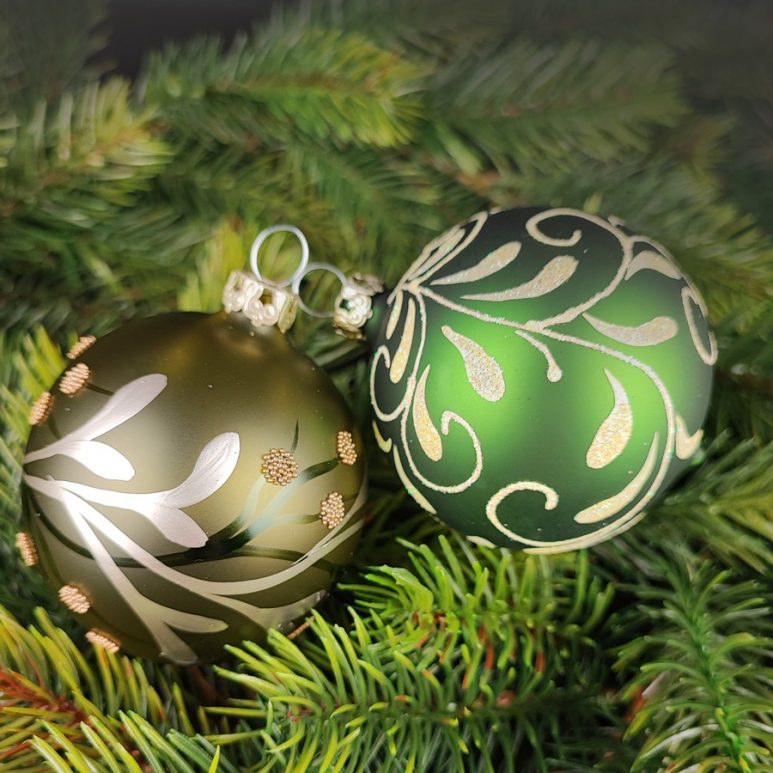 2 Christbaumkugeln aus Glas liegen dekorativ auf grünen Zweigen in einem matten Grün und floralem Design sowie in einem transparentem Khaki-Grün und einer Ranke